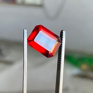 May include: A red gemstone with a rectangular shape is held by tweezers. The gemstone is transparent and has a slight orange hue.