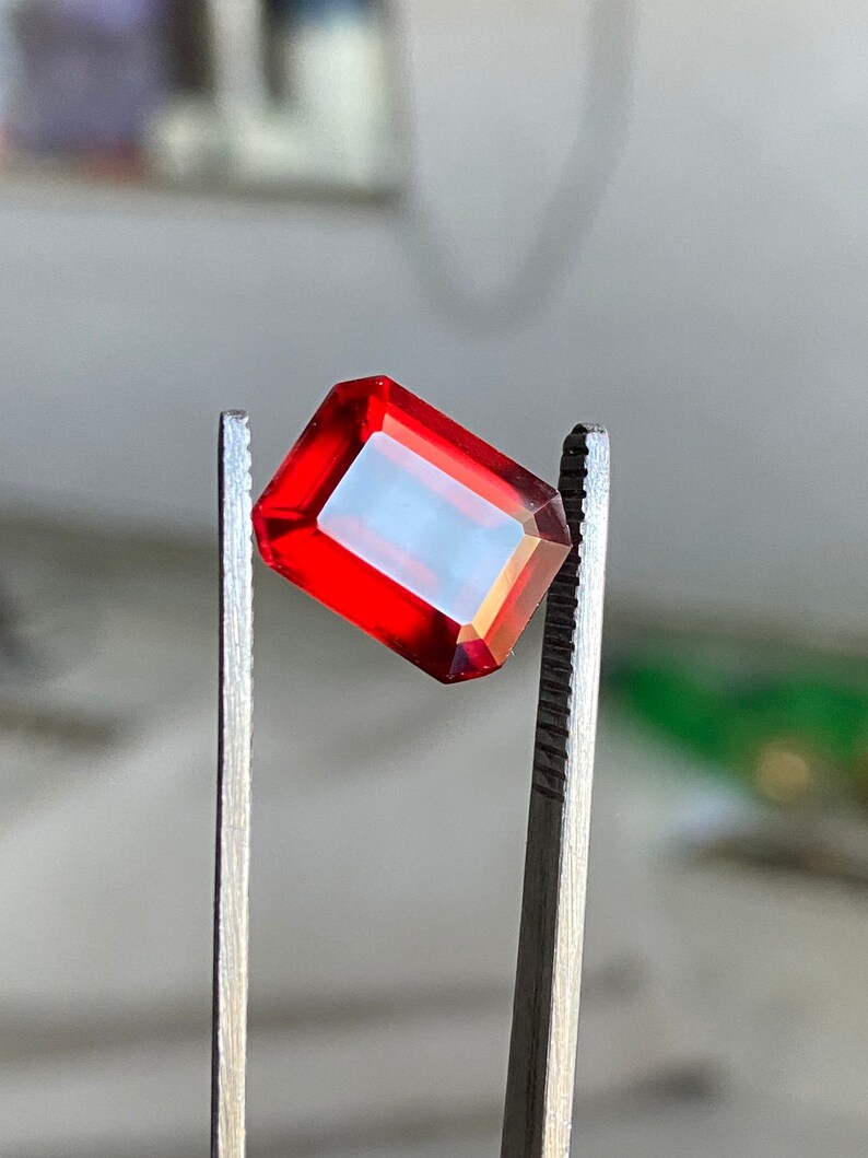 May include: A red gemstone with a rectangular shape is held by tweezers. The gemstone is transparent and has a slight orange hue.