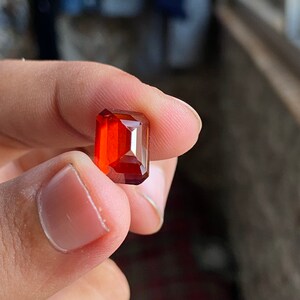 May include: A deep red, rectangular gemstone with a faceted cut. The gemstone is held in a hand.