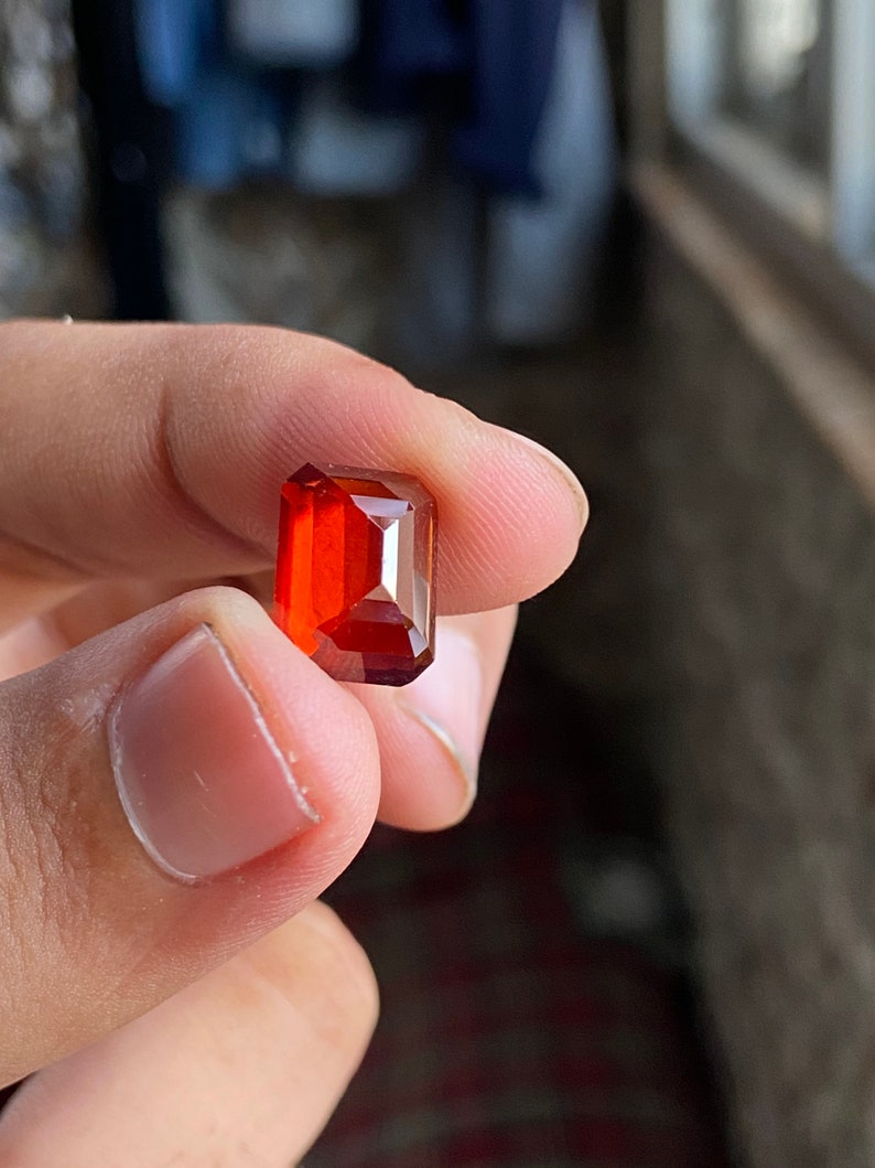 May include: A deep red, rectangular gemstone with a faceted cut. The gemstone is held in a hand.