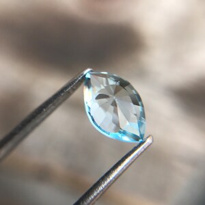 May include: A light blue marquise-shaped gemstone held by tweezers. The gemstone is faceted and has a clear, bright appearance.