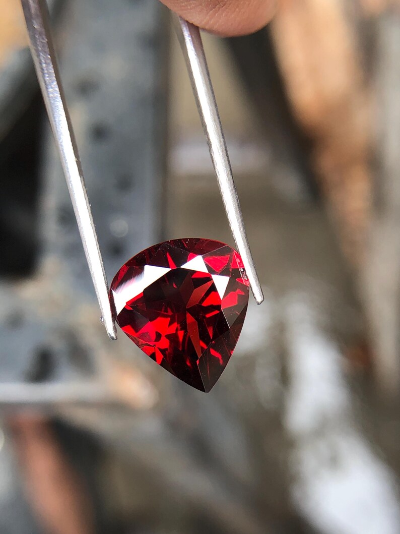May include: A deep red gemstone with a triangular cut, held by tweezers.