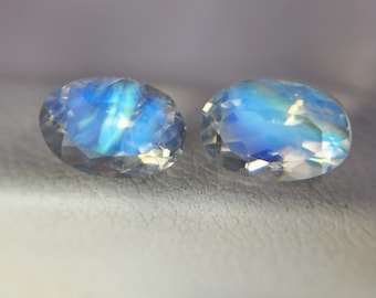 May include: Two oval-shaped, faceted gemstones with a blue and white iridescent glow. The gems have a polished, reflective surface, and are set against a neutral background. The stones appear to be moonstones, known for their adularescence.