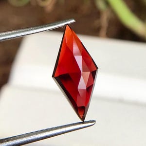 May include: A deep red gemstone with a faceted, kite-shaped cut. The gemstone is held by tweezers.