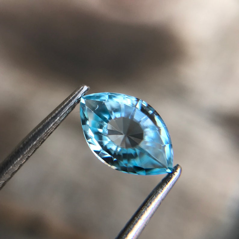 May include: A light blue marquise-shaped gemstone with facets, held by tweezers.