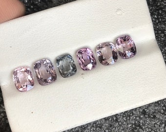 May include: Six cushion-cut gemstones in various shades of pink and gray. The gemstones are arranged in a row on a white surface.