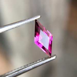 May include: A vibrant pink gemstone with a square shape and a diamond-like cut, held by tweezers.