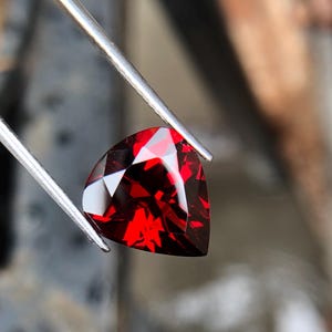 May include: A deep red gemstone with a triangular cut, held by tweezers. The gemstone is faceted and has a high shine.