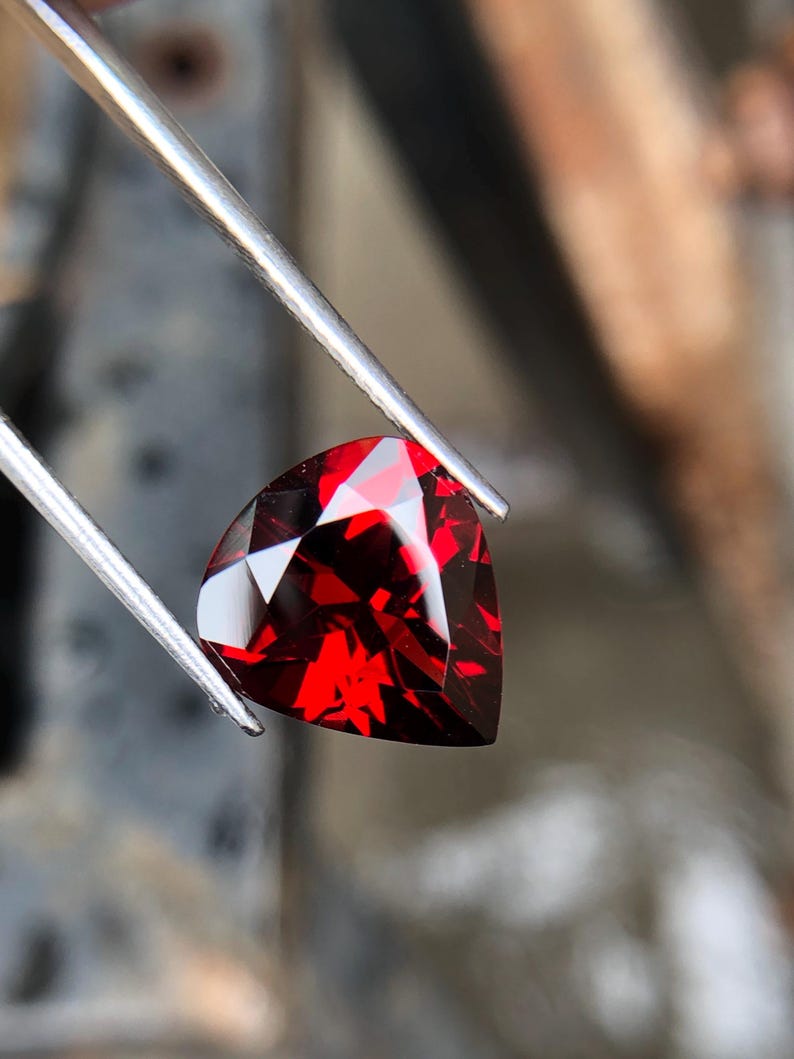 May include: A deep red gemstone with a triangular cut, held by tweezers. The gemstone is faceted and has a high shine.