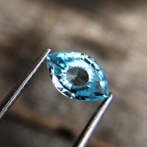 May include: A light blue marquise-shaped gemstone with a faceted design. The gemstone is held in place by a pair of tweezers.