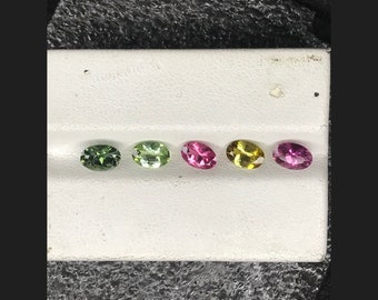 May include: Five oval-shaped gemstones in a row on a white surface. The gemstones are green, green, pink, yellow, and pink.
