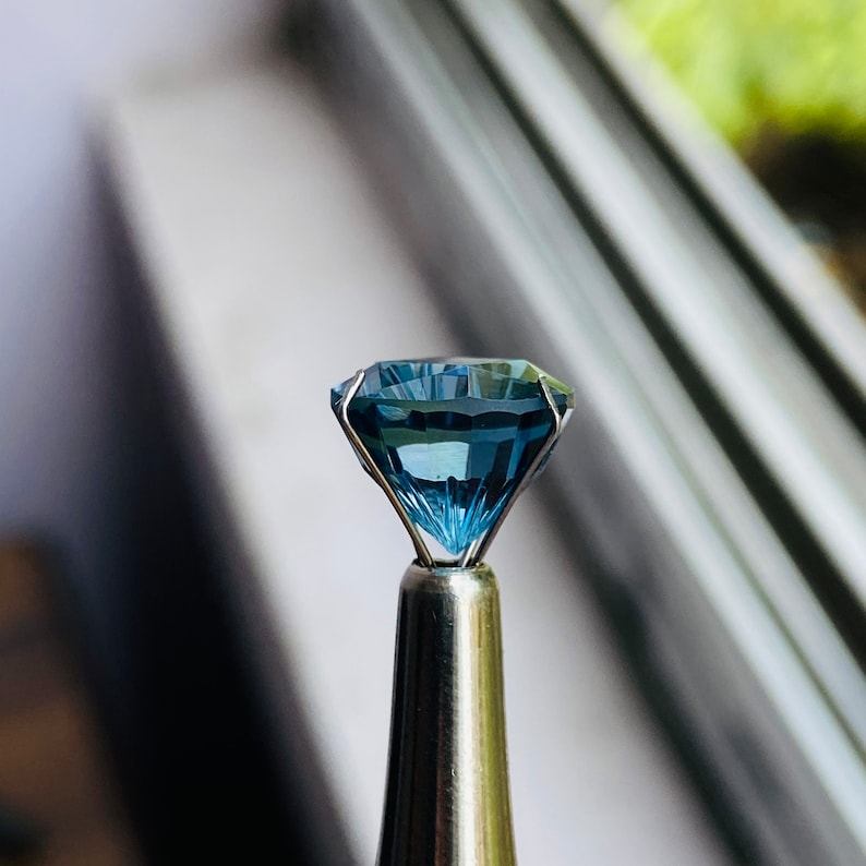 Puede incluir: Una piedra preciosa azul engastada en un metal. La piedra preciosa est&aacute; facetada y tiene un brillo brillante.