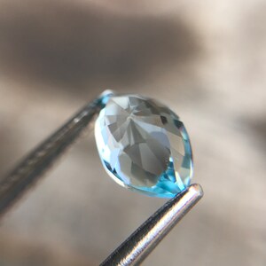 May include: A light blue marquise-shaped gemstone with facets, held by tweezers.