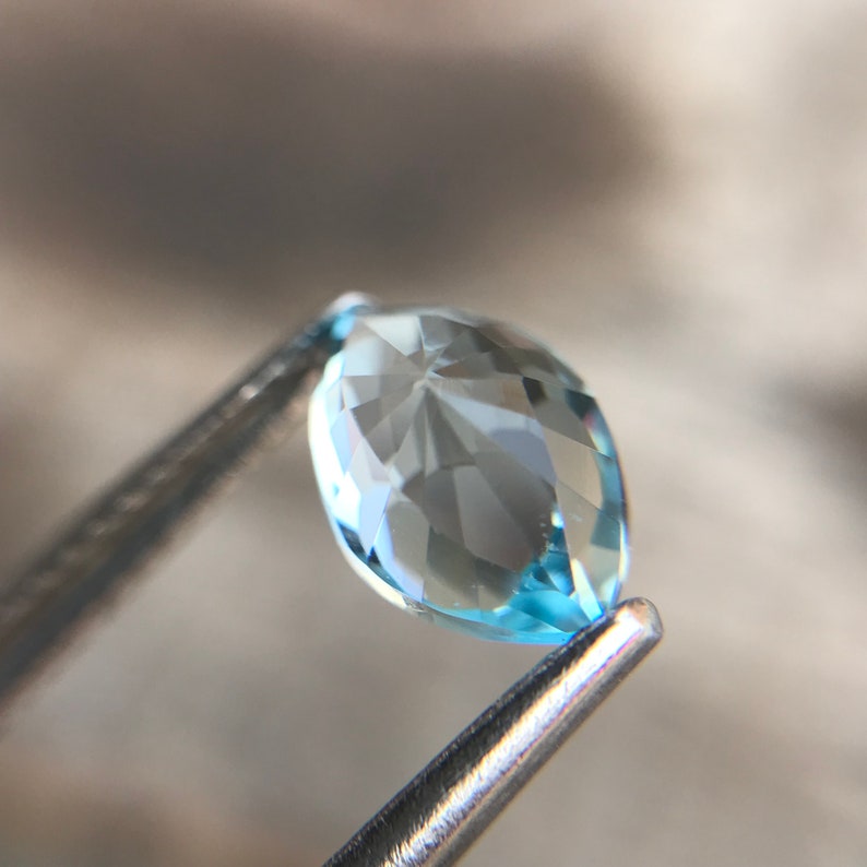 May include: A light blue marquise-shaped gemstone with facets, held by tweezers.