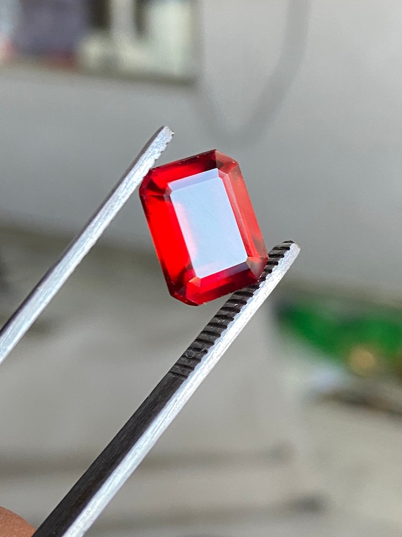 May include: A red gemstone with a rectangular shape is held by a pair of tweezers.