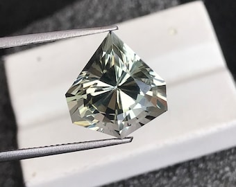 May include: A light green gemstone with a faceted, star-shaped cut. The gemstone is held by tweezers against a white background.