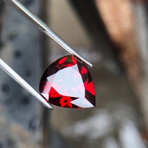 May include: A deep red heart-shaped gemstone with facets, held by tweezers.