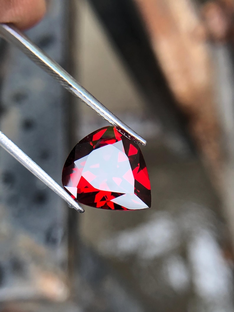 May include: A deep red heart-shaped gemstone with facets, held by tweezers.