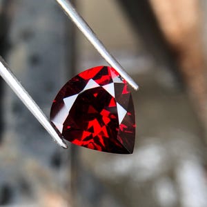 May include: A deep red gemstone with a triangular cut, held by tweezers. The gemstone is faceted and reflects light.