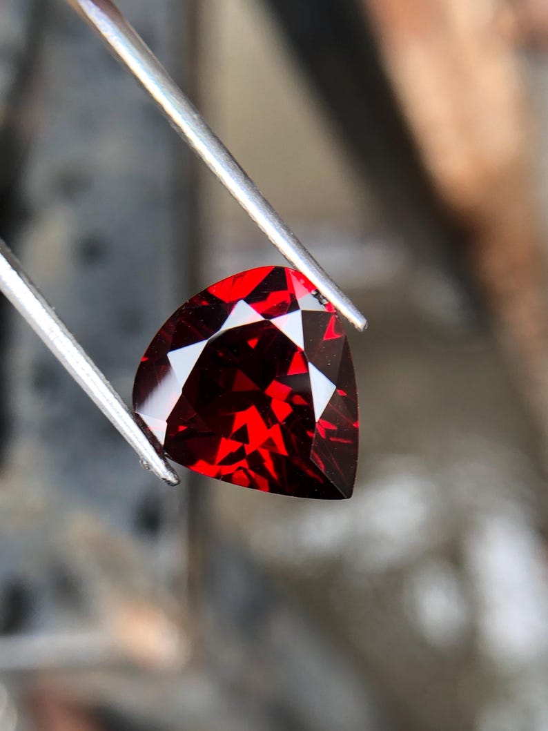 May include: A deep red gemstone with a triangular cut, held by tweezers. The gemstone is faceted and reflects light.