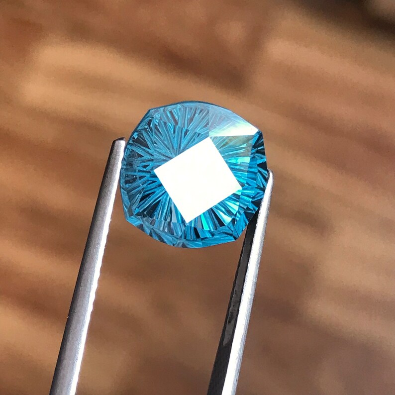 Puede incluir: Una piedra preciosa azul con corte facetado, sostenida por unas pinzas. La piedra preciosa tiene un patr&oacute;n de estrella.