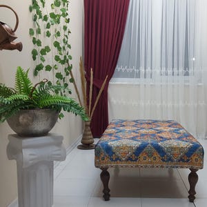 Peut inclure: Un ottoman bleu et or &agrave; motifs avec quatre pieds en bois. L'ottoman est pos&eacute; sur un sol en carrelage blanc dans une pi&egrave;ce aux murs blancs et une fen&ecirc;tre avec des rideaux blancs.