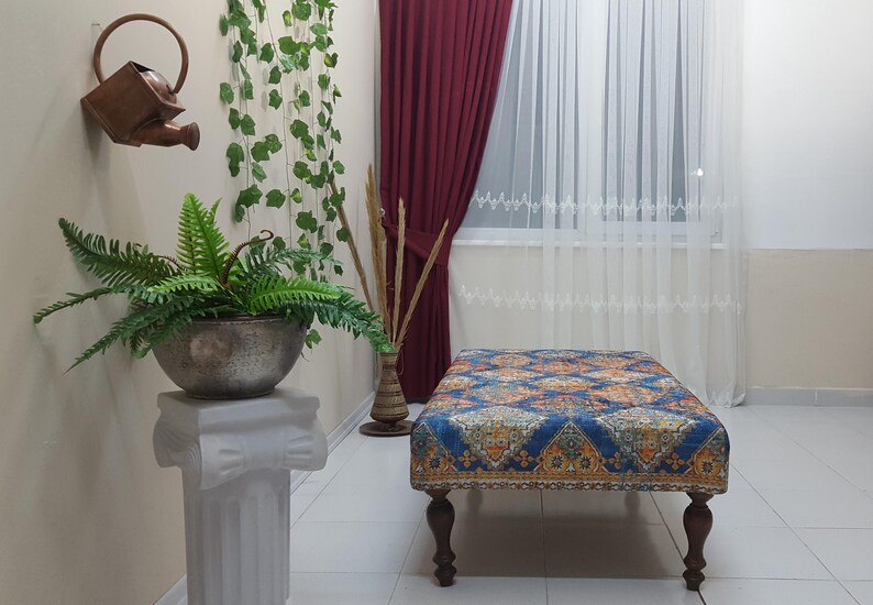 Peut inclure: Un ottoman bleu et or &agrave; motifs avec quatre pieds en bois. L'ottoman est pos&eacute; sur un sol en carrelage blanc dans une pi&egrave;ce aux murs blancs et une fen&ecirc;tre avec des rideaux blancs.