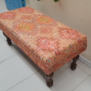 Può includere: Un pouf rettangolare con un top in tessuto a motivi colorati e quattro gambe in legno. Il tessuto presenta un design geometrico in tonalità di rosso, arancione, giallo e bianco.
