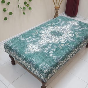 May include: A green and white patterned ottoman with wooden legs. The ottoman has a floral design with a central medallion.