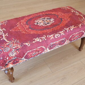 May include: A red and cream patterned ottoman with wooden legs. The ottoman is upholstered in a plush fabric with a traditional design.