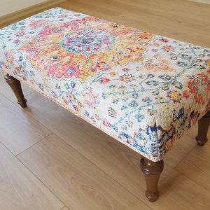 Puede incluir: Un otomano decorativo con un estampado floral colorido y patas de madera. El otomano est&aacute; tapizado en una tela de color crema con un dise&ntilde;o floral multicolor.