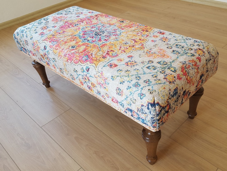 Puede incluir: Un otomano decorativo con un estampado floral colorido y patas de madera. El otomano est&aacute; tapizado en una tela de color crema con un dise&ntilde;o floral multicolor.