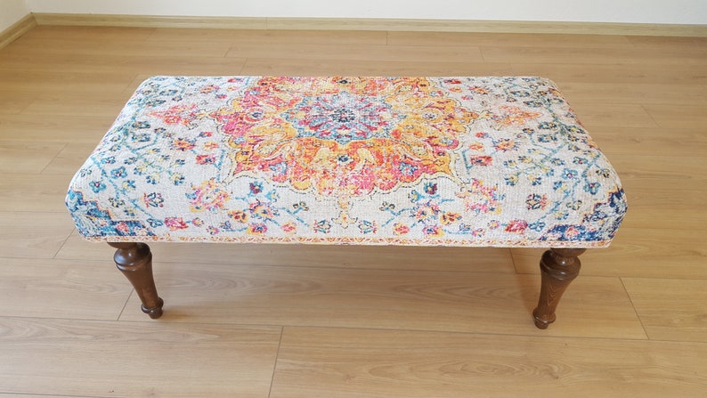 Puede incluir: Un otomano con estampado colorido sobre fondo blanco y patas de madera. El estampado presenta un gran medall&oacute;n central con dise&ntilde;os florales y geom&eacute;tricos en tonos rojos, amarillos, azules y verdes.