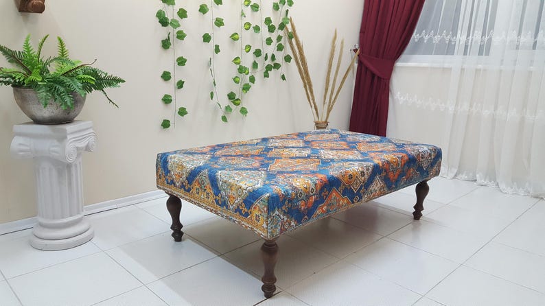 Peut inclure: Un ottoman bleu et orange &agrave; motifs avec des pieds en bois. L'ottoman est pos&eacute; sur un sol en carrelage blanc.