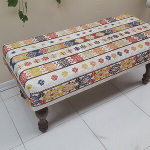 May include: A rectangular ottoman with a white fabric top featuring a colourful geometric pattern. The ottoman has four wooden legs.