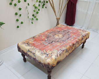 Puede incluir: Un otomano rectangular con una parte superior de tela con estampado floral de colores. El otomano tiene cuatro patas de madera y está sobre un suelo de baldosas blancas.