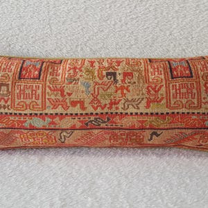 May include: A long, rectangular pillow with a red, orange, and beige patterned fabric. The pattern features geometric shapes and designs.