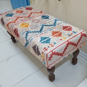 Peut inclure: Un ottoman à motifs avec un fond beige et des motifs géométriques colorés dans des tons de rouge, orange, jaune, bleu et marron. L'ottoman a quatre pieds en bois.