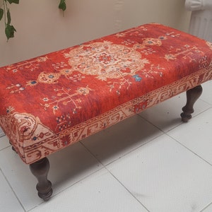 Peut inclure: Un ottoman rouge et crème à motifs avec des pieds en bois foncé. L'ottoman est recouvert d'un tissu qui ressemble à un tapis traditionnel.