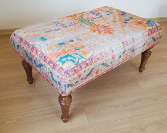Op de afbeelding: Een kleine, rechthoekige poef met een kleurrijk, gepatroneerd stoffen bovenblad en houten poten. De stof heeft een bloemmotief in tinten oranje, roze, blauw en groen.
