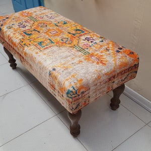 Peut inclure: Un petit ottoman rectangulaire avec un dessus en tissu coloré et à motifs et des pieds en bois. Le tissu présente un motif géométrique dans des tons d'orange, de jaune, de bleu et de blanc.