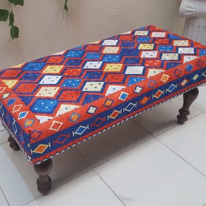 May include: A rectangular ottoman with a colorful geometric pattern in shades of red, blue, yellow, and white. The ottoman has four wooden legs.