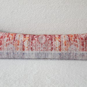 May include: A long, rectangular lumbar pillow with a red, orange, and white patterned fabric. The pattern features a repeating design of stylized flowers and leaves.