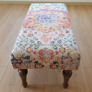 Puede incluir: Un otomano rectangular con una parte superior de tela estampada de colores y patas de madera. La tela presenta un dise&ntilde;o floral en tonos azules, rosas, amarillos y naranjas.