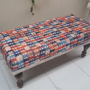 Può includere: Un pouf rettangolare con un motivo geometrico colorato sulla parte superiore. Il pouf ha quattro gambe in legno.