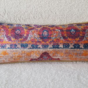 May include: A long, rectangular throw pillow with a colorful, patterned design. The pillow features a red, orange, yellow, and blue geometric pattern reminiscent of a traditional rug.