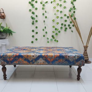Peut inclure: Un ottoman bleu et orange &agrave; motifs avec des pieds en bois. L'ottoman est pos&eacute; sur un sol blanc devant un mur blanc.
