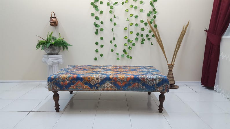 Peut inclure: Un ottoman bleu et orange &agrave; motifs avec des pieds en bois. L'ottoman est pos&eacute; sur un sol blanc devant un mur blanc.
