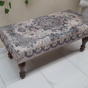 May include: A rectangular ottoman with a beige and gray patterned fabric top and four dark brown wooden legs. The ottoman is sitting on a white tiled floor.