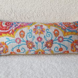May include: A rectangular decorative pillow with a colorful floral pattern. The pillow features a white background with a repeating design of orange, blue, and pink flowers and swirls. The edges are a mix of red, orange, and yellow.